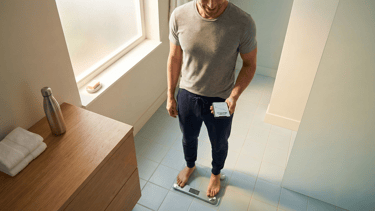 A fit, confident adult in their 30s-40s standing on a modern bathroom scale