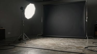 Wide shot of a production setup — a single strobe light aimed at a dark seamless backdrop, cables running across a concrete studio floor, a tripod visible to the left, no people, quiet pre-shoot stillness, high contrast editorial lighting