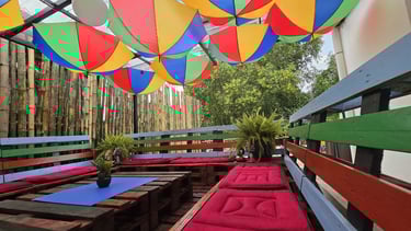 Outdoor patio with pallet seating, red cushions, and colorful umbrellas hanging from the ceiling.