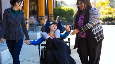 Happy disabled boy in wheelchair outdoors with two smiling female caregivers