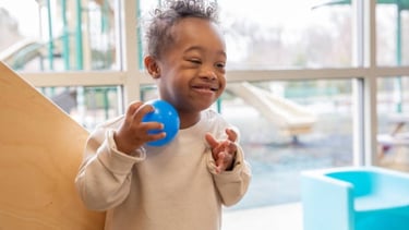Smiling child with Down syndrome playing indoors with a blue ball near window