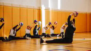 A group of young volleyball players sit on the gym floor holding balls overhead while stretching, le