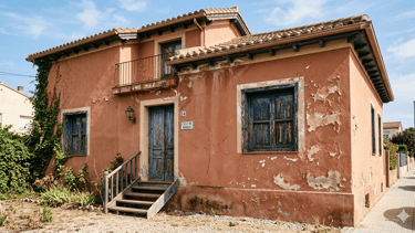 Rustic terracotta Spanish villa with peeling paint, wooden shutters, and a small balcony.