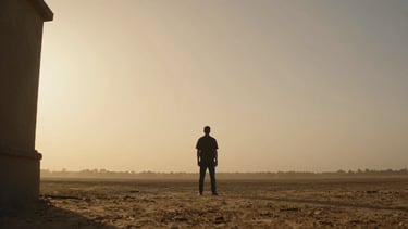 Wide letterbox production still — a lone figure silhouetted against a vast dust-lit exterior location, camera positioned low and wide, golden-hour haze bleeding across the frame, deep shadows framing the left edge, cinematic grain visible