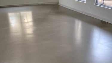 A person is kneeling on the floor in an empty room, placing wooden planks in what seems to be a flooring installation. The room is well-lit by natural light coming through a window. The walls are plain, with a few exposed wires hanging down. There is a bottle placed on the floor, along with several other planks lying around.