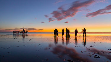 Photographers and horses silhouetted against a vibrant orange sunset on a reflective salt flat water surface.