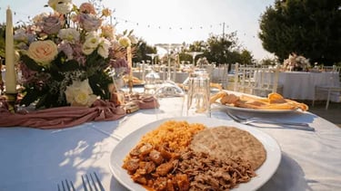 Plato con barbacoa, carnitas, arroz y frijoles sobre mesa elegante con centro de flores en boda al atardecer.
