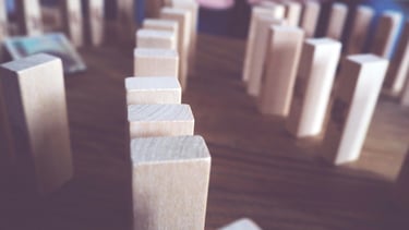 Close-up van houten blokken zoals domino, één valt om, symboliseert gevolgen in de praktijk.