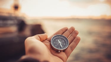 Hand holding a silver pocket compass against a blurred sunset horizon to find his way.