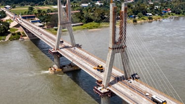 A high-resolution drone photo of a complex bridge construction site over a river in South America. The image shows the technical complexity and precision of the engineering work.