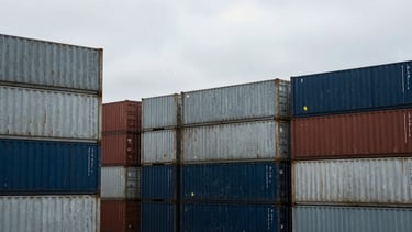 Wide environmental landscape of a massive industrial shipping port under cool overcast daylight, desaturated steel container stacks, sharp focus, 35mm