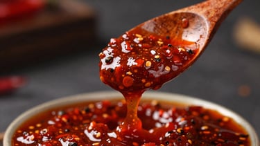 Thick, textured red hot sauce slowly dripping from a rustic wooden spoon, macro shot showing tiny flecks of black pepper and chili seeds, moody lighting.