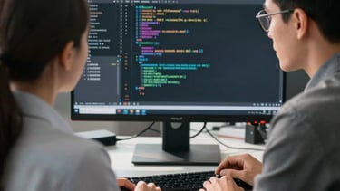 Two professionals in professional attire working together at a large monitor in a sunlit North American office. Sharp focus on the screen showing code, with soft depth of field.