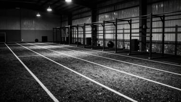 High-contrast monochrome wide shot of an industrial athletic training facility, empty turf lanes, heavy iron racks, dramatic key lights casting deep shadows, raw cinematic grain, 35mm
