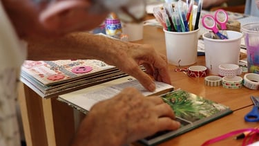 Un adulto mayor, haciendo una manualidad de un libro en una mesa de madera.