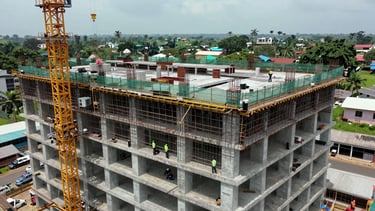 Overhead wide shot of an active high-rise hotel construction site in Nigeria — tower crane visible at left, concrete floor plates at multiple levels under construction, safety netting and formwork in precise arrangement, workers coordinating below in high-visibility vests, natural overcast daylight, documentary aerial framing establishing site scale and operational complexity
