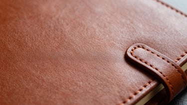 Primer plano macro de un lomo de libreta de piel con costura expuesta, hilo tinto, luz suave enfatizando la textura del cuero.