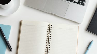 A top-down professional photograph of a minimalist white desk with a laptop, a notebook, and a coffee cup. Soft natural light illuminates the space. Accents of soft blue and light gray are present in the stationery. The style is modern and clean, capturing a Spanish-speaking / Global corporate atmosphere.