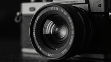Gritty black and white close-up of a 35mm film camera lens reflecting a harsh neon light in a dark room, heavy grain, deep shadows.