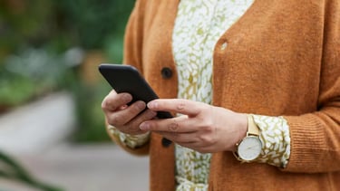 A person in an orange cardigan holding a smartphone in a garden setting.