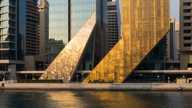 Architectural abstract crop of Dubai waterfront at golden hour, sharp geometric lines of structural glass and brushed brass, casting long dramatic shadows across a calm harbor, panoramic 21:9 composition.