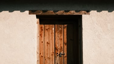 Tight abstract crop of a weathered teak door frame set against raw textured plaster, harsh high-contrast sunlight casting deep shadows, 35mm film.