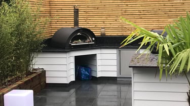 An outdoor barbecue area. A pizza oven is fixed in the corner in front of a wooden slatted fence..