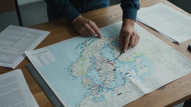 Overhead close study of a large planning surface, a hand-annotated map of the North Atlantic with city markers, a ruler, printed documents and handwritten notes scattered across a wooden table, cool diffused studio light, no digital screens