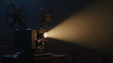 Atmospheric low-key film still of a vintage movie projector casting a warm golden light beam through a dark room, rich navy shadows, high-contrast chiaroscuro, 35mm grain.