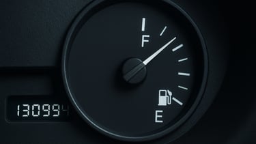 Extreme close-up overhead shot of a car dashboard odometer and fuel gauge mid-reading, illuminated by cool white studio light against a dark charcoal surface, technical instrument detail visible, no human faces