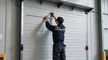 A detailed professional photograph of a technician installing a heavy-duty industrial roll-up door in a North American warehouse. The lighting is bright and industrial, highlighting the metallic textures of the silver door and the dark navy work gear of the professional. The composition is a medium shot focusing on the precision of the installation, conveying reliability and modern expertise.