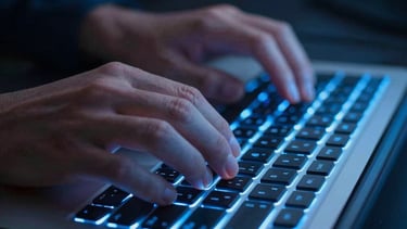 A close-up photograph of hands typing on a high-end backlit keyboard in a dimly lit North American office. The blue glow of the keys matches the deep navy and sky blue color palette.