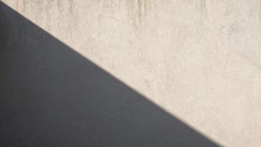 Wide bleed image of a textured plaster wall under harsh direct sunlight, creating a deep, sharp geometric shadow.