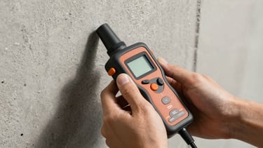Close-up of hands holding a digital acoustic leak detector against a concrete wall, high-contrast daylight.