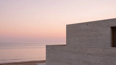 Wide panoramic shot of a minimalist concrete villa overlooking a calm sea at sunset, warm pink sky, soft architectural shadows, 35mm photography