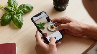 A top-down professional photography shot in a sunlit European / Spanish studio. A person is arranging high-end gastronomic photos, a smartphone showing social media feeds, and fresh basil leaves on a light beige wooden desk. The composition is clean and elegant, featuring accents of crimson red and forest green. Soft natural lighting creates a warm, approachable atmosphere.
