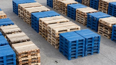 A high-angle, professional photograph of an organized North American industrial yard. Neatly stacked wooden and blue plastic pallets are arranged in rows under bright daylight. The scene emphasizes clean operations and modern pallet management efficiency with muted blue and light grey tones.