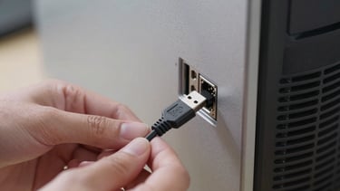 Close-up shot of hands connecting a USB peripheral cable into the rear I/O panel of a desktop tower, clinical bright ambient lighting, steel gray case visible, cable and port detail sharp and clear, no moody shadows