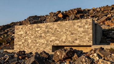 Wide-angle view of a brutalist stone facade integrated into the topography of a rocky hillside, evening golden hour.