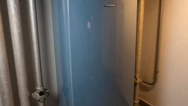 Professional high-angle shot of a modern, clean mechanical room with insulated pipes and a high-efficiency boiler, warm lighting reflecting off metallic surfaces, Est European / Român architectural interior style, featuring colors like Slate Blue and Tan.
