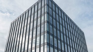 A sharp, wide-angle architectural photograph of a modern commercial office building in Ilford, United Kingdom. The image features a clean glass facade reflecting a light grey and medium blue sky, conveying a sense of transparency and modern professionalism. The lighting is bright and clear, emphasizing reliability.