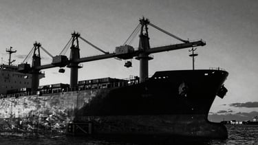 Low-key black and white editorial photography of a massive cargo ship hull and industrial cranes at dusk, dramatic chiaroscuro lighting, deep shadows, sharp contrast, 35mm film grain