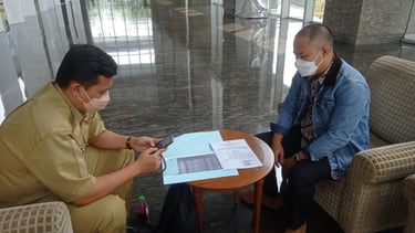 Two men wearing face masks conduct a formal business meeting with documents in a modern building lobby.