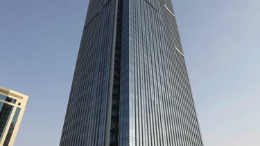 A professional wide-angle architectural photograph of a sleek glass office tower in the Ajman Free Zone. The building reflects a clear sky, featuring sophisticated shades of dark navy and silver. The scene is set in a modern Middle Eastern / Gulf commercial district, conveying a sense of authority and global reach through clean lines and bright, natural sunlight.