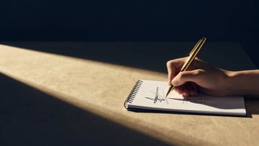 Wide cinematic shot of a minimalist, sunlit Parisian office, a single designer's hand sketching on a notepad with a gold pen, soft dramatic studio light, deep navy and gold tones, high-contrast, Hasselblad, wide aspect