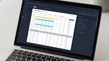Wide-angle overhead shot of a dark-navy ERP dashboard on a laptop screen, showing real-time analytics charts, payroll summary cards, and inventory counts — office desk with keyboard partially visible, bright studio lighting, tight on-screen crop