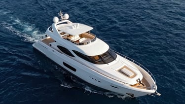 Aerial view of a sleek, modern yacht on the Mediterranean sea, shot from a drone. The water is a deep steel blue, the boat a bright pearl white.