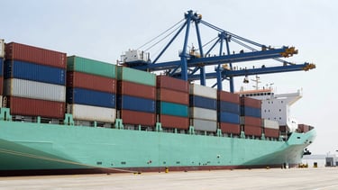 Wide-angle industrial logistics port with stacked shipping containers and a cargo vessel docked, bright clean daylight, sharp focus.