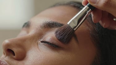 Extreme close-up panoramic strip of a makeup brush pressing pigment onto an eyelid on medium brown skin, soft diffused window light revealing every hair of the brush and the texture of the lid, hands steady mid-stroke, warm neutral background