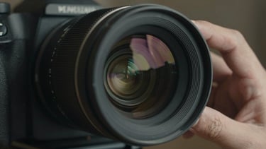 Extreme close-up of a high-end cinema camera lens reflecting dramatic studio lights, hands adjusting the focus ring, shallow depth of field, moody charcoal tones.